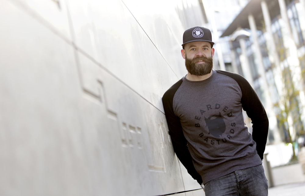 man wearing black fitted cap, black-and-gray raglan crew-neck long-sleeved shirt and denim bottoms leaning at wall during daytime