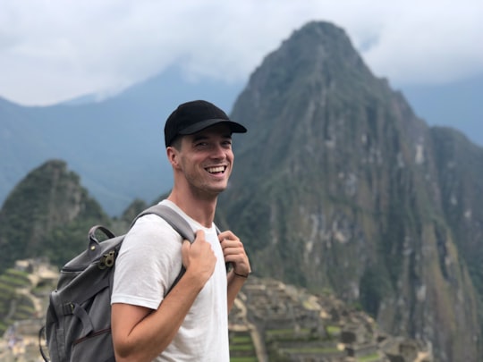 smiling man with backpack stands on crest facing green mountain in Machu Picchu Peru