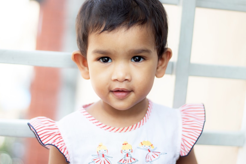 A young child with short dark hair is wearing a white dress with red and white striped ruffle sleeves and colorful embroidery of dancers on the front. The child is standing outdoors in front of a background featuring a soft focus pattern of vertical and horizontal lines.