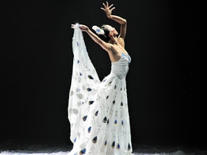 A dancer gracefully poses with arms extended above her head, wearing a flowing white dress adorned with peacock feather patterns set against a dark background. The lighting highlights the elegant lines and movement.