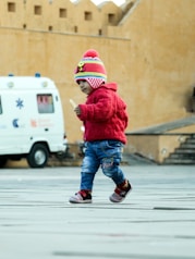A young child wearing a colorful knit hat and a red sweater is walking across a paved area. The child seems to be holding a small object in their hand. In the background, there is a white van with medical symbols and some text. The setting appears to be an outdoor area with large, beige-colored walls.