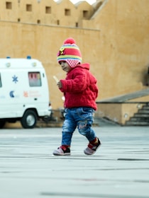 A young child wearing a colorful knit hat and a red sweater is walking across a paved area. The child seems to be holding a small object in their hand. In the background, there is a white van with medical symbols and some text. The setting appears to be an outdoor area with large, beige-colored walls.