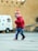 A young child wearing a colorful knit hat and a red sweater is walking across a paved area. The child seems to be holding a small object in their hand. In the background, there is a white van with medical symbols and some text. The setting appears to be an outdoor area with large, beige-colored walls.