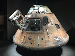 A vintage spacecraft capsule with a conical shape, featuring an open hatch revealing mechanical details inside. The capsule has a worn, metallic exterior with visible bolts and panels, indicating past use in space exploration. It is displayed in a dimly lit environment, suggesting a museum or exhibit setting.