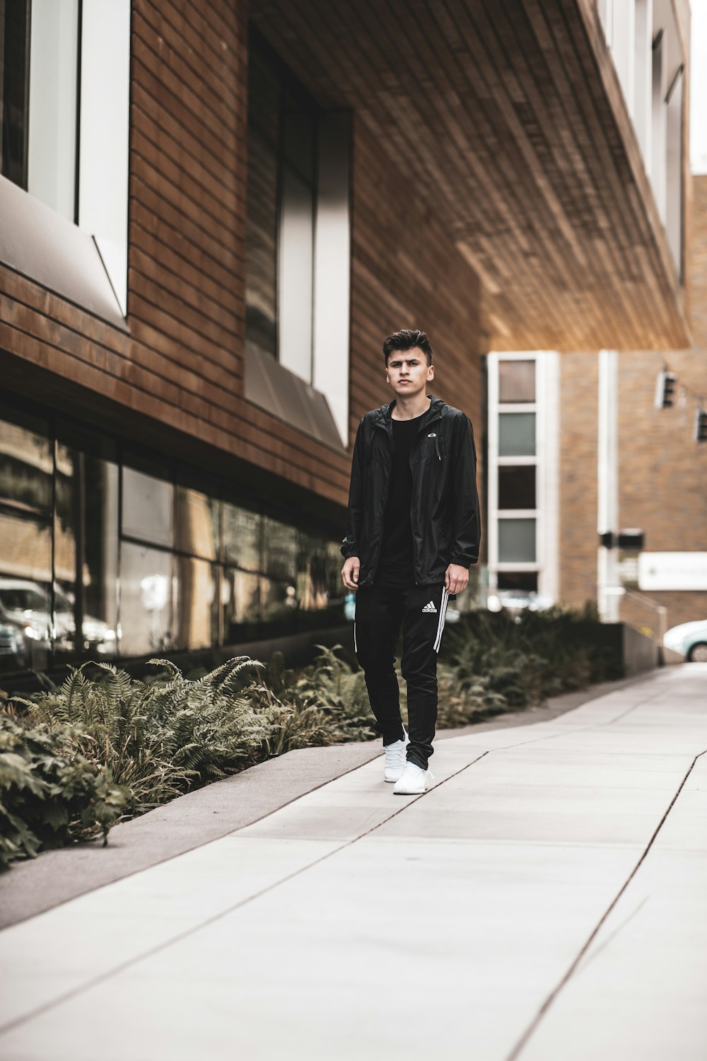 man wearing black leather zip jacket, black shirt and black pants outfit standing beside brown building