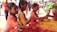 Three young children are playing enthusiastically with vibrant colored powders, covering themselves and their surroundings in hues of red, yellow, and orange. Their expressions are joyful and engaged as they explore the textures and colors. Several other people are visible in the blurred background, suggesting a festive and communal celebration.