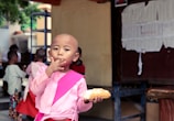 A child wearing a pink outfit is eating a bread roll and using their hand to touch their lips. The background shows several other children, some with shaved heads, standing and engaged in various activities. A wall with papers pinned on it is visible to the right.