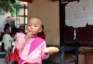 A child wearing a pink outfit is eating a bread roll and using their hand to touch their lips. The background shows several other children, some with shaved heads, standing and engaged in various activities. A wall with papers pinned on it is visible to the right.