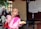 A child wearing a pink outfit is eating a bread roll and using their hand to touch their lips. The background shows several other children, some with shaved heads, standing and engaged in various activities. A wall with papers pinned on it is visible to the right.