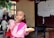 A child wearing a pink outfit is eating a bread roll and using their hand to touch their lips. The background shows several other children, some with shaved heads, standing and engaged in various activities. A wall with papers pinned on it is visible to the right.