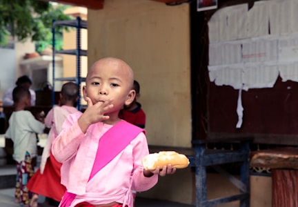 A child wearing a pink outfit is eating a bread roll and using their hand to touch their lips. The background shows several other children, some with shaved heads, standing and engaged in various activities. A wall with papers pinned on it is visible to the right.