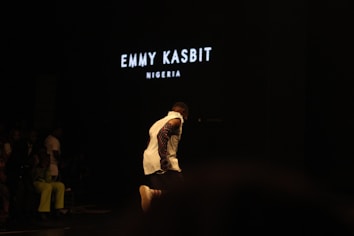 A person is walking on a dark stage, wearing a white top with patterned sleeves. The background is dimly lit with the words 'EMMY KASBIT NIGERIA' prominently displayed in white. A few other people are faintly visible on the side, seemingly seated and watching.