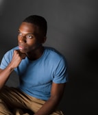 A person sits thoughtfully, resting their chin on their hand. They are wearing a light blue t-shirt and khaki pants. The background is gray with soft lighting focused on the person's face, creating a contemplative and introspective atmosphere.