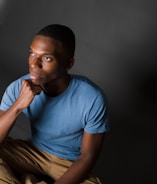 A person sits thoughtfully, resting their chin on their hand. They are wearing a light blue t-shirt and khaki pants. The background is gray with soft lighting focused on the person's face, creating a contemplative and introspective atmosphere.