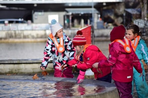 Children dressed warmly in colorful winter clothing are gathered around a stone edge near water. They appear to be playing or exploring, with one child reaching out towards the water. Orange safety vests are visible on their outfits.