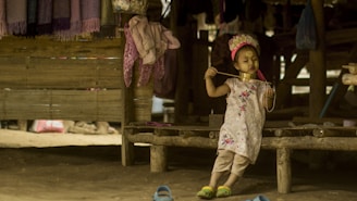 A young child, adorned with traditional neck rings and face paint, stands on a wooden platform in a rustic setting. The child appears to be wearing a decorative headpiece and a floral dress. Surrounding the child are hanging textiles and casually placed items, creating an atmospheric, cultural scene.