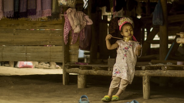 A young child, adorned with traditional neck rings and face paint, stands on a wooden platform in a rustic setting. The child appears to be wearing a decorative headpiece and a floral dress. Surrounding the child are hanging textiles and casually placed items, creating an atmospheric, cultural scene.