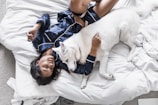 A person wearing dark blue silk pajamas is lying on a bed covered with white sheets, cuddling with a large white dog. The person appears relaxed and at ease, and the dog looks calm and content. The surrounding environment includes a portion of a white cushion and an unmade bed.