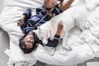 A person wearing dark blue silk pajamas is lying on a bed covered with white sheets, cuddling with a large white dog. The person appears relaxed and at ease, and the dog looks calm and content. The surrounding environment includes a portion of a white cushion and an unmade bed.