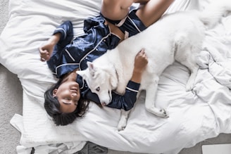 A person wearing dark blue silk pajamas is lying on a bed covered with white sheets, cuddling with a large white dog. The person appears relaxed and at ease, and the dog looks calm and content. The surrounding environment includes a portion of a white cushion and an unmade bed.