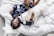 A person wearing dark blue silk pajamas is lying on a bed covered with white sheets, cuddling with a large white dog. The person appears relaxed and at ease, and the dog looks calm and content. The surrounding environment includes a portion of a white cushion and an unmade bed.