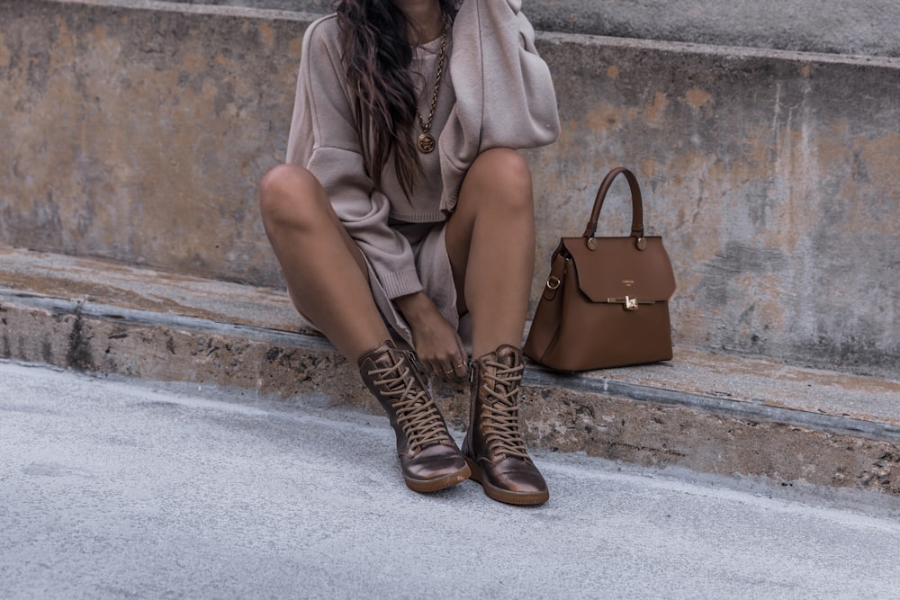Mujer sentada en una superficie de hormigón marrón con botas marrones al lado de un bolso de cuero marrón