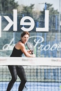 A woman playing padel, holding a paddle with both hands, dressed in athletic wear on a court. A net and a glass wall with partially visible text are present. The background shows some trees and a clear sky.