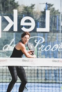 A woman playing padel, holding a paddle with both hands, dressed in athletic wear on a court. A net and a glass wall with partially visible text are present. The background shows some trees and a clear sky.