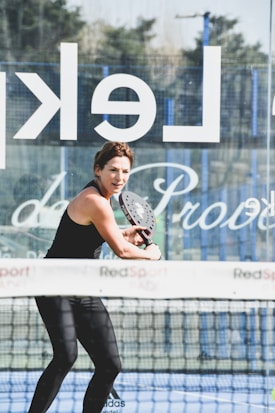 A woman playing padel, holding a paddle with both hands, dressed in athletic wear on a court. A net and a glass wall with partially visible text are present. The background shows some trees and a clear sky.
