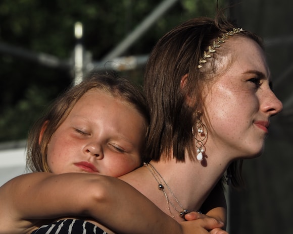 A young girl, eyes closed, rests her head on the shoulder of a woman wearing a beaded headband and earrings. The woman gazes forward with a serene expression, and the atmosphere suggests a moment of calm and closeness.