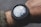 A digital watch displaying GPS coordinates and altitude readings is worn on a person's wrist. The watch face shows detailed numerical data against a dark, rugged design. The person's arm is clothed in a green, textured fabric, possibly an outdoor jacket.