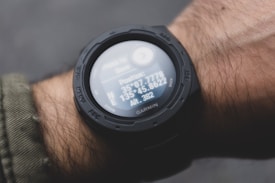 A digital watch displaying GPS coordinates and altitude readings is worn on a person's wrist. The watch face shows detailed numerical data against a dark, rugged design. The person's arm is clothed in a green, textured fabric, possibly an outdoor jacket.