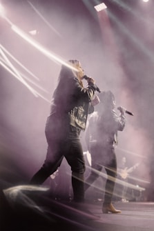 Two people are performing on stage, holding microphones. They are surrounded by dramatic lighting and smoke effects, wearing dark clothing. The background includes a guitarist and beams of light cutting through the haze.