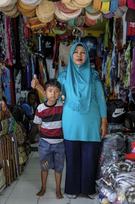 A woman in a turquoise hijab and blue dress stands beside a young boy in a striped shirt and plaid shorts. They are inside a shop filled with colorful clothes and woven hats hanging overhead.