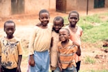 Five children are standing closely together outdoors, smiling and posing for the photo. The setting appears to be a rural area with a dirt ground and simple buildings in the background. The children wear casual clothing, and one has a shirt featuring a graphic of a wolf.