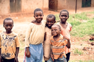 Five children are standing closely together outdoors, smiling and posing for the photo. The setting appears to be a rural area with a dirt ground and simple buildings in the background. The children wear casual clothing, and one has a shirt featuring a graphic of a wolf.