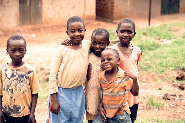 Five children are standing closely together outdoors, smiling and posing for the photo. The setting appears to be a rural area with a dirt ground and simple buildings in the background. The children wear casual clothing, and one has a shirt featuring a graphic of a wolf.