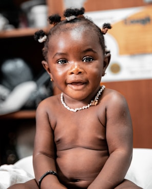 A young child with short hair styled in small puffs is sitting and smiling, wearing a pearl necklace and a black bracelet. The background is slightly blurred, indicating an indoor environment, possibly a room with shelves.