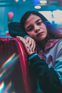 A young person leans against a shiny red surface, appearing thoughtful. The lighting is soft with colorful reflections, adding a dreamy effect. The background is blurred with cool blue tones, emphasizing the subject's relaxed posture and subtle expression.