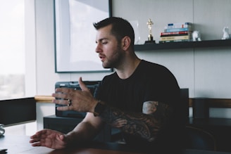 A man with tattoos on his arm is sitting at a desk in an office, gesturing as he speaks. Shelves in the background hold books and a small statue.