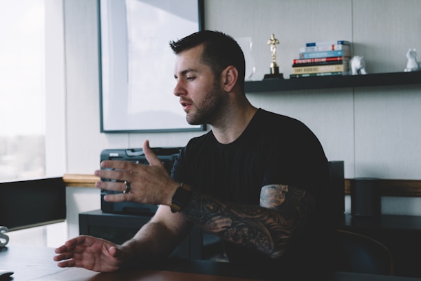 A man with tattoos on his arm is sitting at a desk in an office, gesturing as he speaks. Shelves in the background hold books and a small statue.
