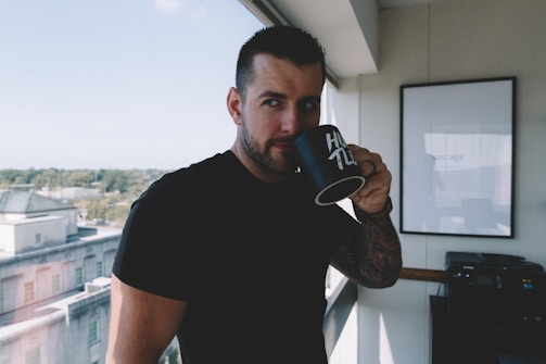 A person with short hair is standing indoors, sipping from a black mug with the word 'HUSTLE' printed on it. They are next to a window overlooking a cityscape and the sky is clear. Inside, there is a framed picture on the wall and a printer nearby.