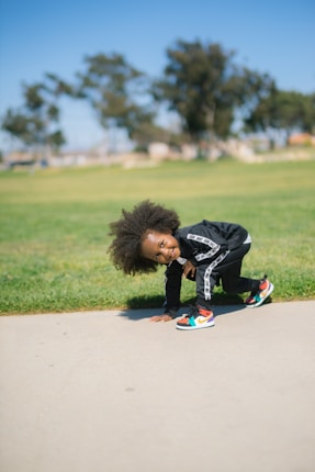 A child with curly hair is playfully bending over on a paved path in a park. They are wearing a black tracksuit and colorful sneakers, with green grass and blurred trees in the background on a sunny day.