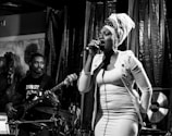 A woman with a headwrap sings passionately into a microphone on stage, while a man plays a bass guitar beside her. The lighting creates a dramatic atmosphere with deep shadows and highlights.