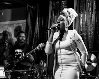 A woman with a headwrap sings passionately into a microphone on stage, while a man plays a bass guitar beside her. The lighting creates a dramatic atmosphere with deep shadows and highlights.
