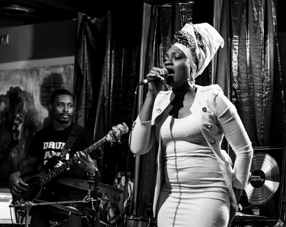 A woman with a headwrap sings passionately into a microphone on stage, while a man plays a bass guitar beside her. The lighting creates a dramatic atmosphere with deep shadows and highlights.