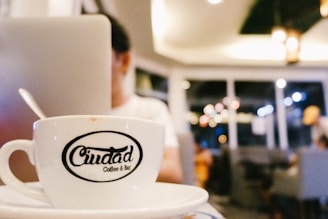 A close-up view of a coffee cup with the logo 'Ciudad Coffee & Bar' on it, placed on a table in a modern cafe setting. The background is softly blurred, showing a person sitting with a laptop and warm ambient lighting with bokeh effects from hanging lights.