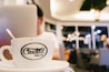 A close-up view of a coffee cup with the logo 'Ciudad Coffee & Bar' on it, placed on a table in a modern cafe setting. The background is softly blurred, showing a person sitting with a laptop and warm ambient lighting with bokeh effects from hanging lights.
