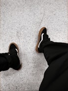 Black sneakers with white accents are worn on a speckled, light gray floor. The shoes have brown soles, and the person is wearing dark pants.