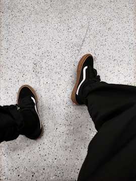 Black sneakers with white accents are worn on a speckled, light gray floor. The shoes have brown soles, and the person is wearing dark pants.